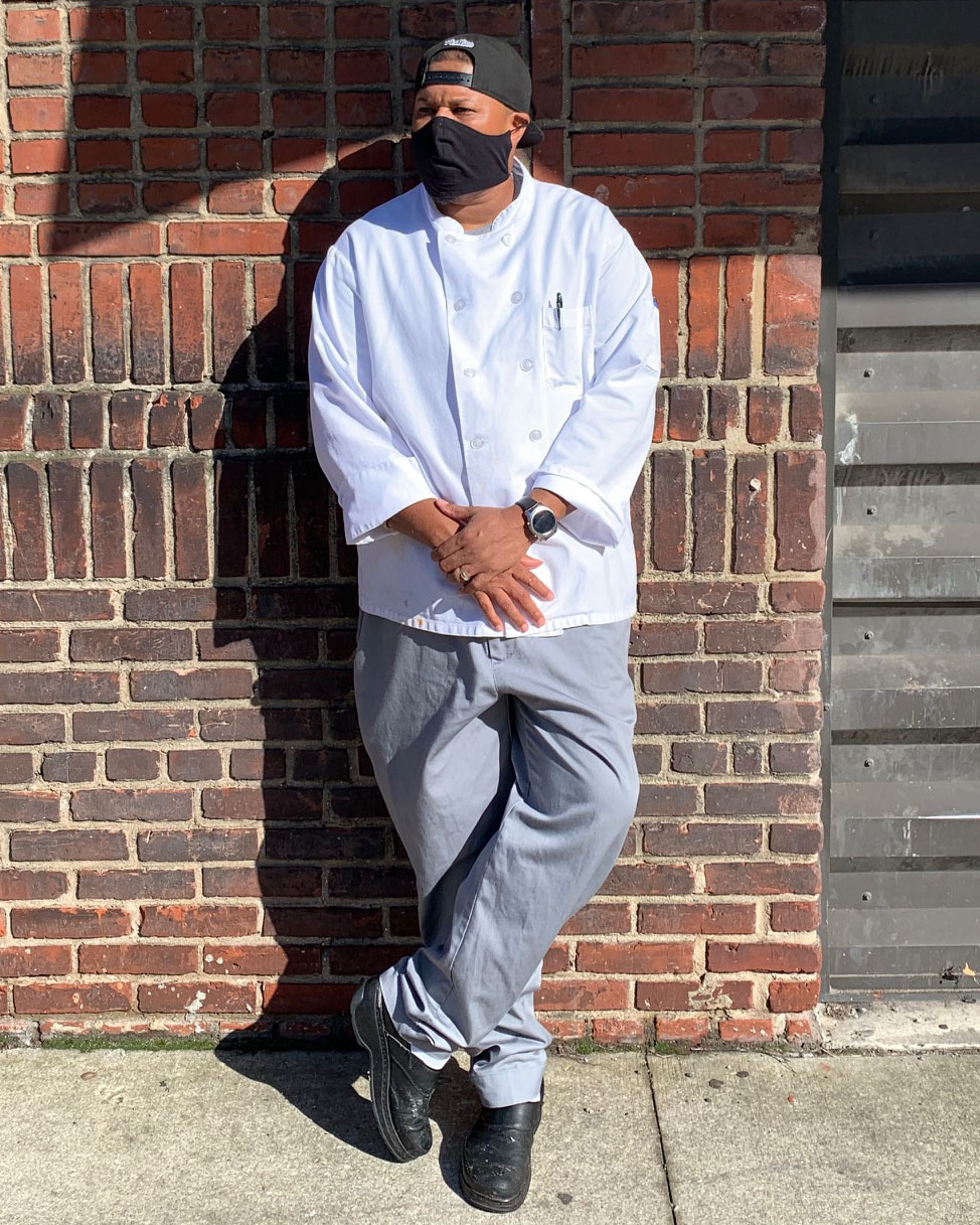 black man in a chef coat and face mask leaning on a wall