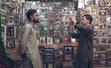 man filming an interview with a second man in a room with photos covering the walls