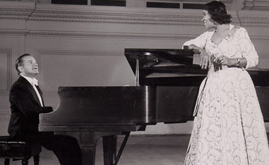 black and white photo woman standing next to piano