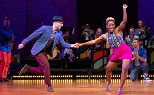 a man and a women swing dancing on a stage with backup dancers